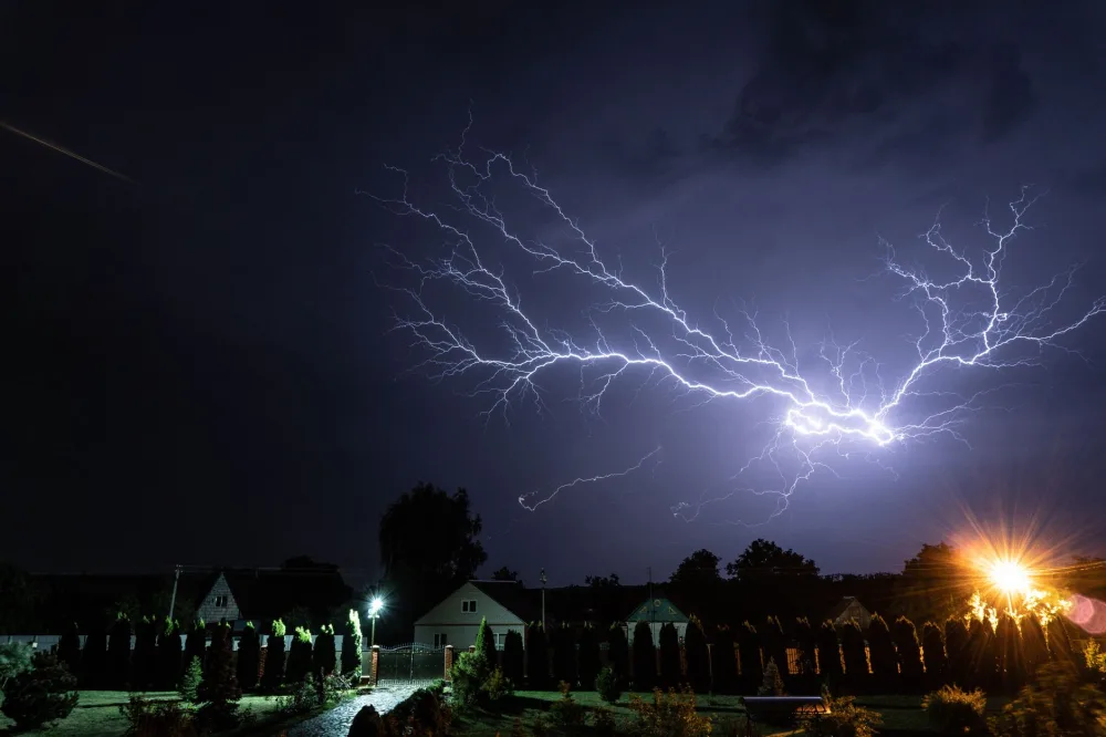 A powerful lightning bolt illuminates the night sky above a suburban neighborhood during a thunderstorm.
