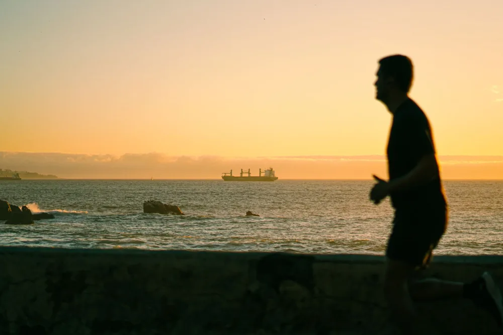 A silhouette of a jogger running along the oceanfront at sunset, with a ship on the horizon.