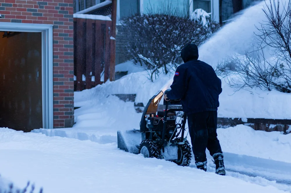 A person using a snow blower to clear snow from a suburban sidewalk in Toronto during winter.