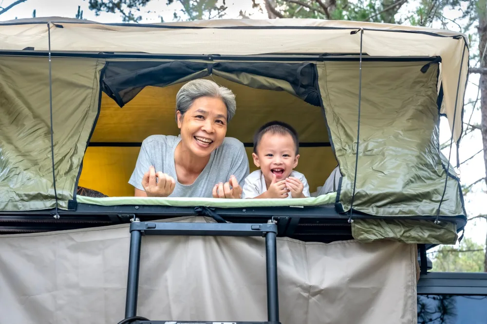 Smiling grandmother with child in a rooftop tent during a joyful camping trip.